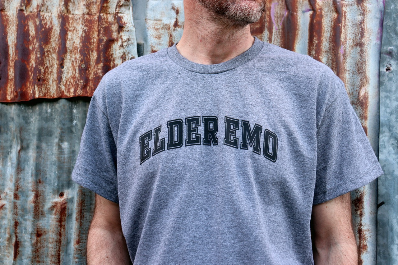 Person wearing a gray t-shirt with 'ELDER EMO' text against a rusty metal wall.