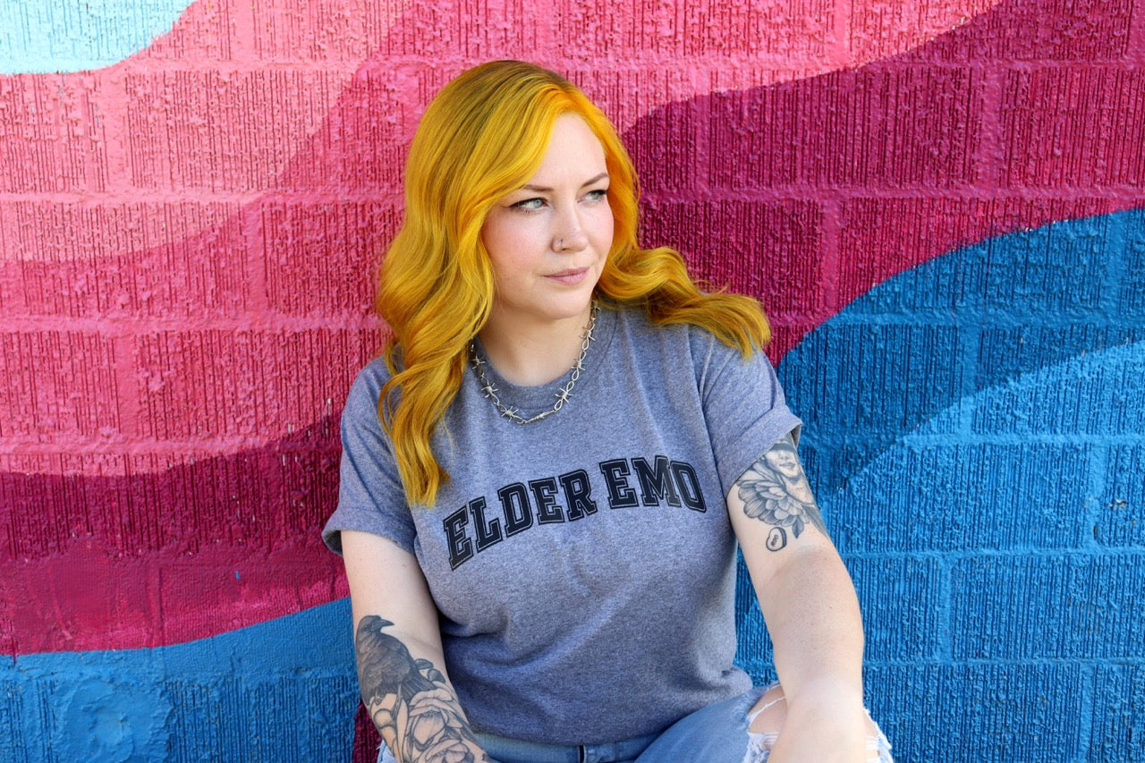 Person with colorful hair and tattoos wearing a 'Elder Emo' shirt against a multicolored textured wall.