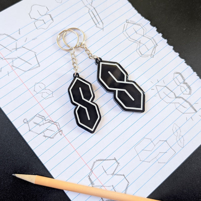 Black geometric keychains on a piece of paper with drawings and a pencil.