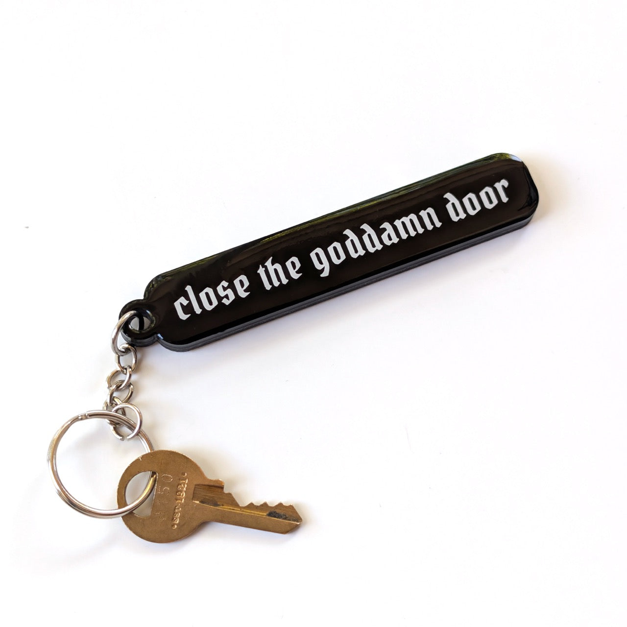 A black rubber keychain with the phrase 'close the goddamn door' in white gothic-style lettering, attached to a silver metal keyring with a brass key, shown on a plain white background.