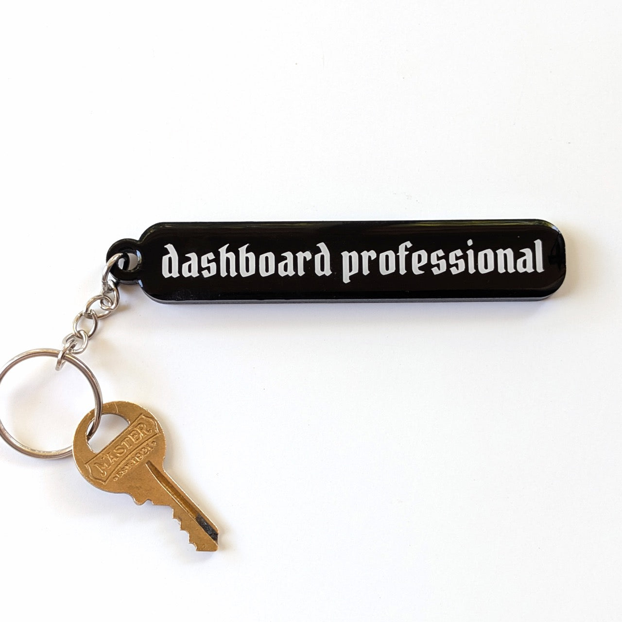 A black rectangular keychain with the words 'dashboard professional' in white gothic-style lettering, attached to a silver keyring with a brass key, on a plain white background.